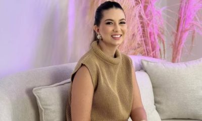 Na imagem, vemos uma mulher jovem sentada em um sofá bege claro. Ela está usando uma blusa de tricô sem mangas de cor bege e calças claras