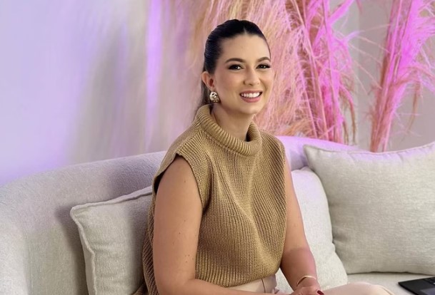 Na imagem, vemos uma mulher jovem sentada em um sofá bege claro. Ela está usando uma blusa de tricô sem mangas de cor bege e calças claras