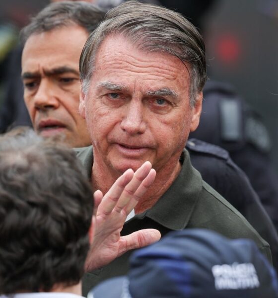 Jair Bolsonaro cercado por pessoas durante evento, olhando para a câmera com expressão séria