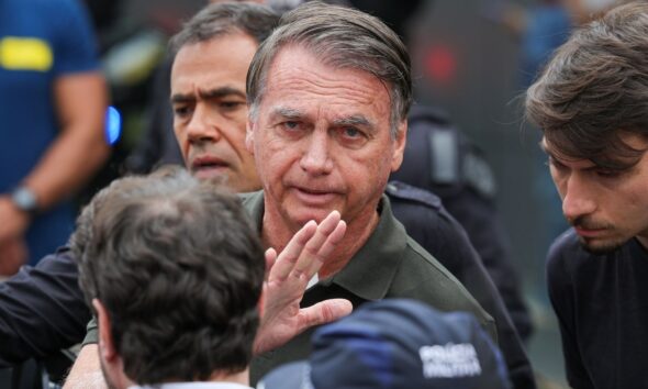 Jair Bolsonaro cercado por pessoas durante evento, olhando para a câmera com expressão séria