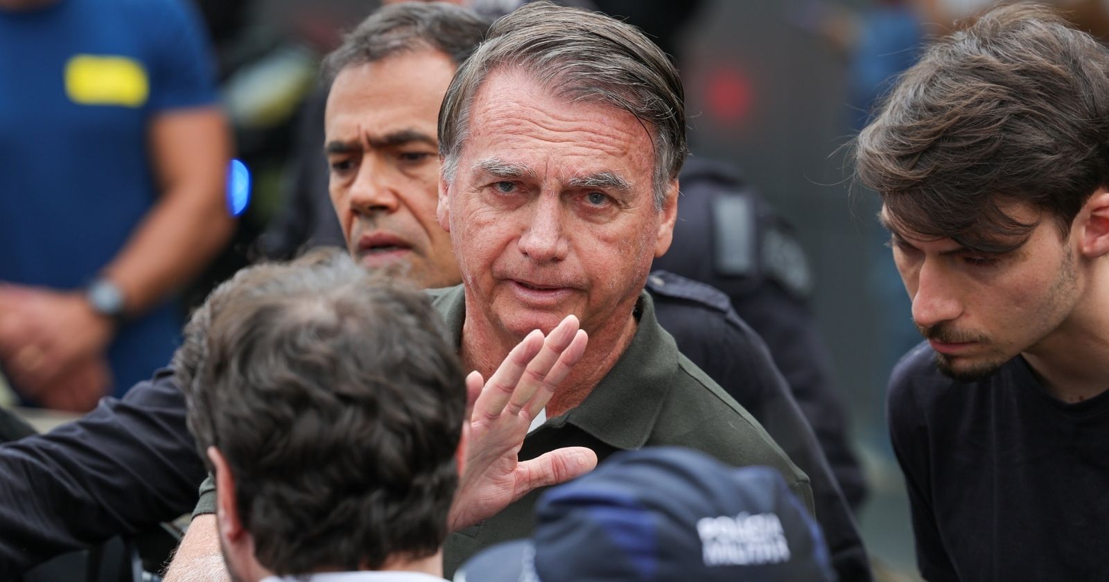 Jair Bolsonaro cercado por pessoas durante evento, olhando para a câmera com expressão séria