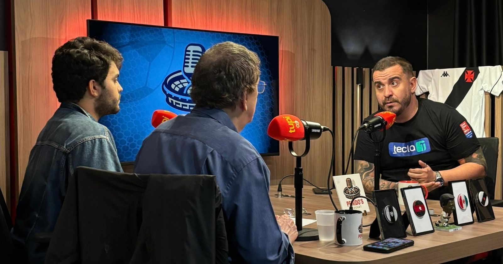 FuiClear sentado à mesa do estúdio da Rádio Tupi durante gravação do Penido's Podcast com Rafa e Luiz Penido