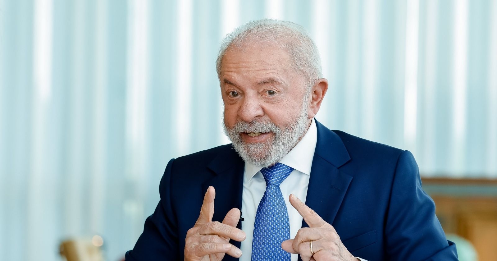 Presidente Lula de terno azul e gravata azul clara, gesticulando com as mãos durante fala em ambiente interno com fundo claro
