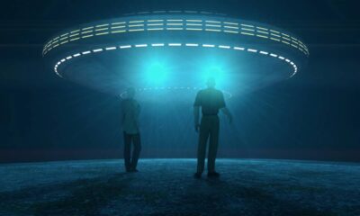 Ilustração mostra silhuetas humanas observando objeto voador não identificado com luzes azuis no céu noturno, representando busca por vida extraterrestre