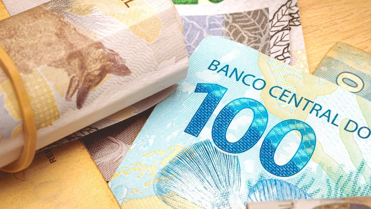 Cédula brasileira de cem reais em destaque, com maço de notas enroladas e outras cédulas.