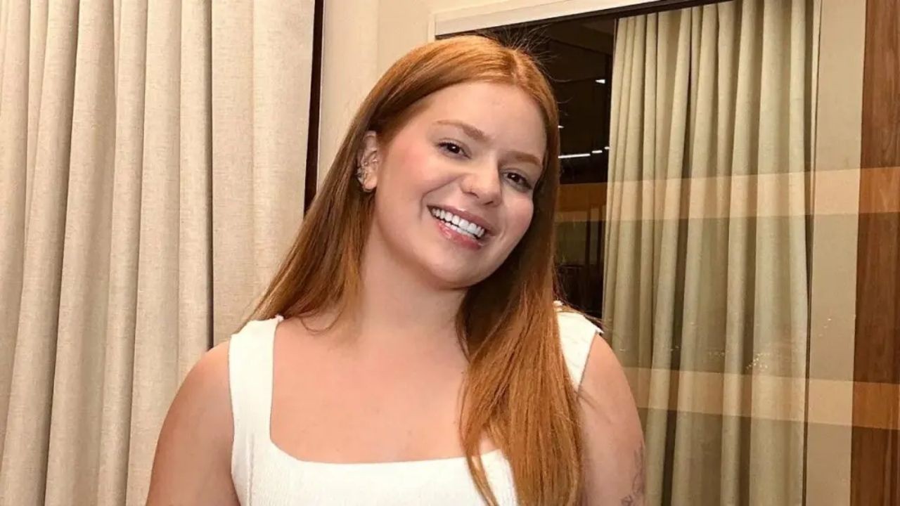 Mulher ruiva de cabelos longos e blusa branca sorri para a câmera, com cortinas ao fundo.