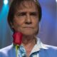 Roberto Carlos, com expressão séria, segura rosas vermelhas em palco azul-iluminado.