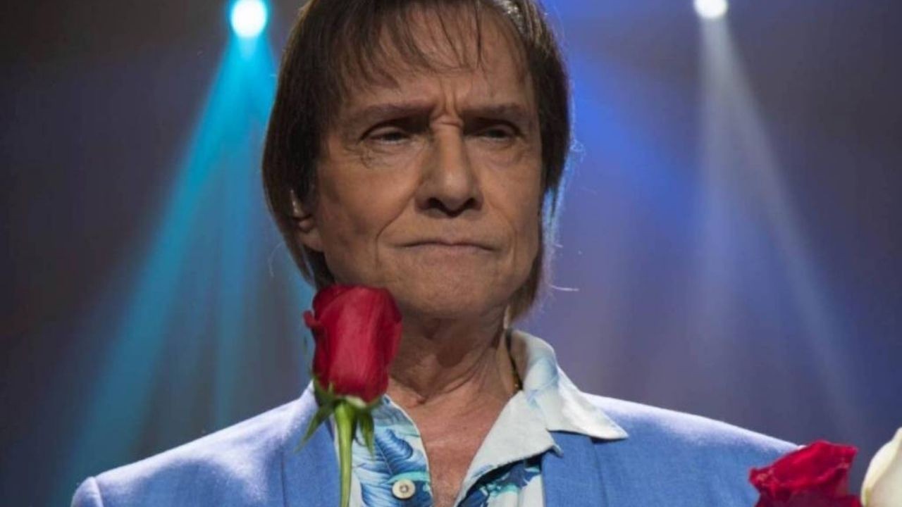 Roberto Carlos, com expressão séria, segura rosas vermelhas em palco azul-iluminado.