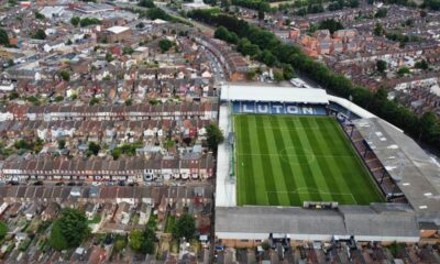 Vista aérea de estádio de futebol com "LUTON" nas arquibancadas, cercado por casas urbanas.