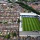 Vista aérea de estádio de futebol com "LUTON" nas arquibancadas, cercado por casas urbanas.