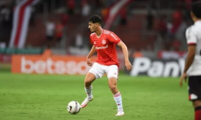 Jogador de futebol em camisa vermelha e shorts brancos, driblando bola em campo verde.