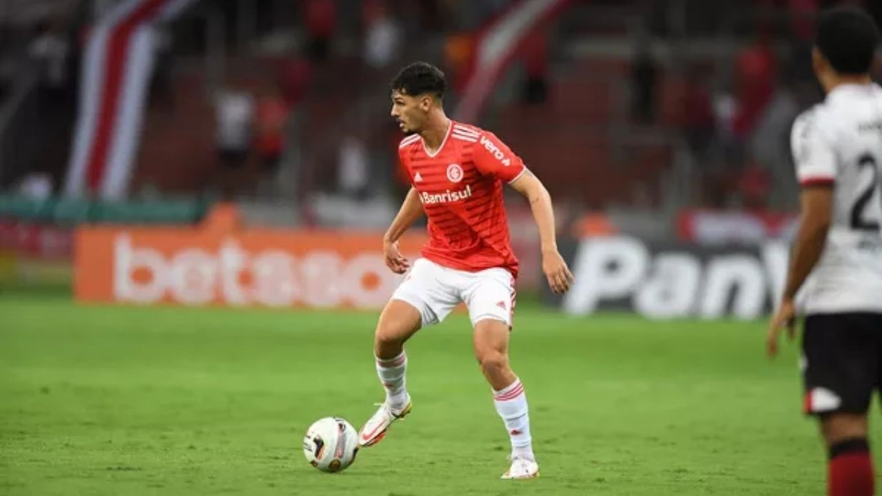 Jogador de futebol em camisa vermelha e shorts brancos, driblando bola em campo verde.