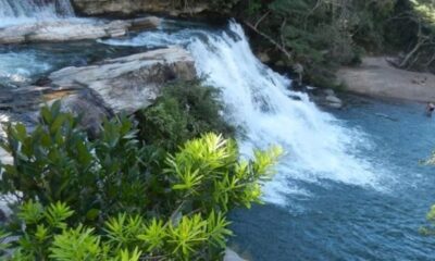 Cachoeira em rio azul, rochas, vegetação verde, pessoas na água e areia.