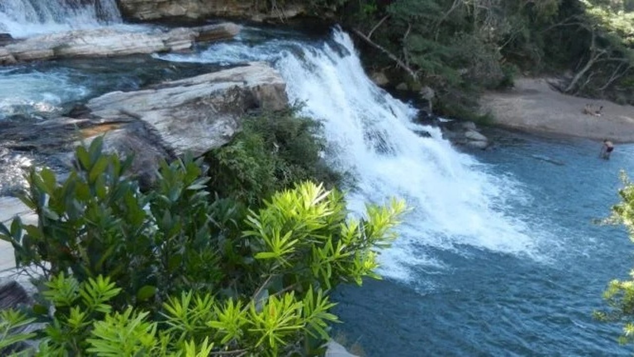 Cachoeira em rio azul, rochas, vegetação verde, pessoas na água e areia.