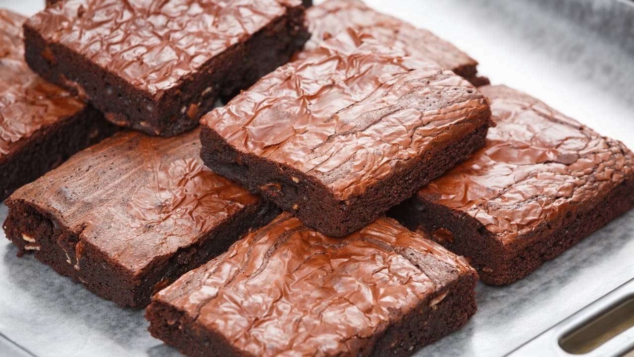 Vários brownies de chocolate escuro com topo brilhante e crocante sobre papel manteiga.