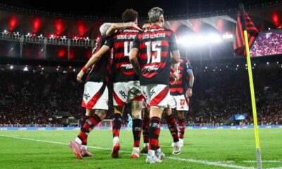 Jogadores de futebol comemoram em campo, vestindo camisas rubro-negras, números 4 e 15 visíveis.