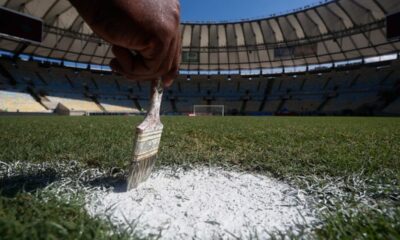 Mão pinta linha branca em campo de futebol com estádio e gol ao fundo.