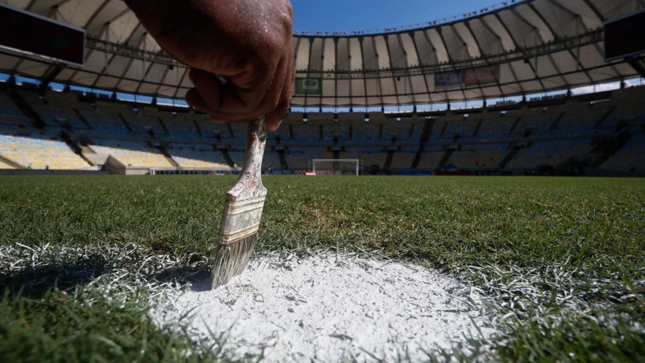 Mão pinta linha branca em campo de futebol com estádio e gol ao fundo.
