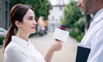 Mulher entrevistando homem com microfone branco, ele segura um caderno, ao ar livre.