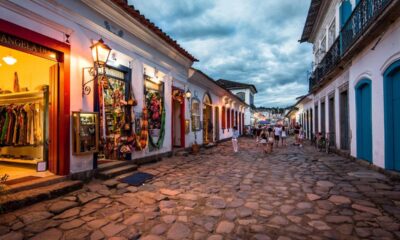 Rua de paralelepípedos ladeada por casas coloniais brancas iluminadas ao entardecer, com lojas e pessoas.