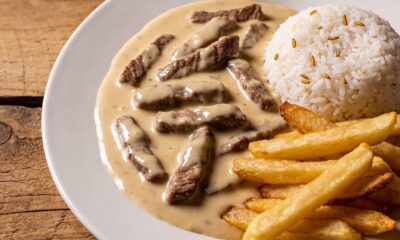 Entre os pratos mais presentes na mesa do brasileiro, o estrogonofe de carne ocupa lugar fixo. A combinação de tirinhas de carne macia com um molho encorpado e cremoso costuma agradar diferentes paladares e se adapta bem tanto ao dia a dia quanto a ocasiões especiais, com ingredientes simples e preparo rápido, sem exigir técnicas complexas de cozinha. O que torna o estrogonofe de carne clássico tão popular A popularidade do estrogonofe de carne clássico se explica, em grande parte, pela combinação de praticidade e sabor marcante. A receita utiliza cortes de carne acessíveis, como alcatra ou chã de dentro, e permite variações sem perder a essência, mantendo um custo relativamente baixo. Outro ponto que contribui é a facilidade de servir, pois o prato harmoniza com acompanhamentos simples, o que reduz o tempo de preparo da refeição completa. Além disso, a textura suave agrada tanto crianças quanto adultos, ajudando a manter a receita presente nas cozinhas domésticas e em cardápios de restaurantes. Como fazer estrogonofe de carne cremoso passo a passo Para preparar um estrogonofe de carne cremoso, o primeiro cuidado está na escolha e no corte da carne. Cortes como filé mignon, alcatra ou chã de dentro costumam garantir maciez quando fatiados em tirinhas finas, facilitando o cozimento rápido e uniforme. Em seguida, entra a etapa de fritura das tirinhas em pequenas porções, em óleo bem quente, para dourar a superfície e manter o interior macio. Assim, a carne não solta líquido em excesso, preservando a textura, o sabor e a cremosidade do molho que será preparado na mesma panela. Ver essa foto no Instagram Uma publicação compartilhada por Arthur Paek (@arthurpaek) Quais são os ingredientes do estrogonofe de carne clássico A receita tradicional de estrogonofe de carne se baseia em poucos ingredientes, mas a combinação entre eles gera um molho equilibrado. A carne bem escolhida e temperada se une ao creme de leite, ao ketchup, ao extrato de tomate e à mostarda, resultando em um prato marcante. Para facilitar o preparo, a seguir está uma versão estruturada da receita com os itens principais utilizados na carne e no molho cremoso. Essa organização ajuda a separar as etapas e torna o processo mais prático na hora de cozinhar. Carne em tiras (filé mignon, alcatra ou chã de dentro), temperada com sal, pimenta e cominho; Cebola ralada para o refogado e óleo quanto baste para fritar as tiras; Creme de leite, ketchup, extrato de tomate e mostarda para formar o molho; Ajustes opcionais de tempero, como pimenta-do-reino ou ervas frescas, conforme o gosto. Ingredientes para a carne 400 g de carne em tiras (filé mignon, alcatra ou chã de dentro) Sal a gosto Pimenta a gosto Cominho a gosto 1/2 cebola ralada Óleo quanto baste para fritar Ingredientes para o molho cremoso 1 caixa e meia de creme de leite 3 colheres (sopa) de ketchup 3 colheres (sopa) de extrato de tomate 1 colher (sopa) de mostarda Modo de preparo detalhado Preparar e temperar a carne Cortar a carne em tirinhas de tamanho semelhante para garantir cozimento por igual. Temperar com sal, pimenta e cominho, misturando bem para envolver todas as tiras. Deixar descansar por cerca de 20 minutos, em local refrigerado, para potencializar o sabor. Dourar as tirinhas Aquecer óleo suficiente em uma panela larga, em fogo médio a alto. Colocar de 7 a 8 tirinhas por vez, evitando amontoar, e dourar rapidamente. Retirar cada leva assim que a carne estiver levemente dourada, para não ressecar. Reservar todas as tiras fritas em um recipiente separado. Preparar a base do molho Descartar o excesso de óleo da panela e deixar apenas uma fina camada. Adicionar a cebola ralada e refogar em fogo médio, mexendo, até dourar. Voltar todas as tirinhas de carne para a panela, misturando com a cebola. Finalizar o molho cremoso Acrescentar o creme de leite, o ketchup, o extrato de tomate e a mostarda. Misturar bem até formar um molho uniforme, envolvendo toda a carne. Deixar ferver em fogo baixo por alguns minutos, mexendo, até atingir a cremosidade desejada. O guia completo do estrogonofe de carne ideal para famílias - Créditos: (depositphotos.com / ws-studio) Como servir estrogonofe de carne de maneira equilibrada O modo de servir o estrogonofe tradicional influencia diretamente na experiência à mesa e no equilíbrio nutricional do prato. A combinação mais comum inclui arroz branco e batata palha, em porções moderadas, para evitar que o prato fique pesado em excesso. Para tornar a refeição mais completa, algumas pessoas acrescentam saladas simples, como folhas verdes ou legumes cozidos, que trazem contraste de textura e cor. Em almoços em família, o estrogonofe costuma ser servido em travessas aquecidas, facilitando que cada pessoa se sirva conforme a preferência, seja em refeições do dia a dia ou em ocasiões especiais em 2025.