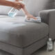 Pessoa aplicando álcool com amaciante em sofá de tecido, método popular para limpeza leve e manutenção do cheiro agradável.