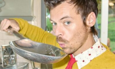 Harry Styles com suéter amarelo sopra uma colher de metal, em ambiente interno.