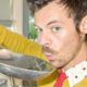 Harry Styles com suéter amarelo sopra uma colher de metal, em ambiente interno.