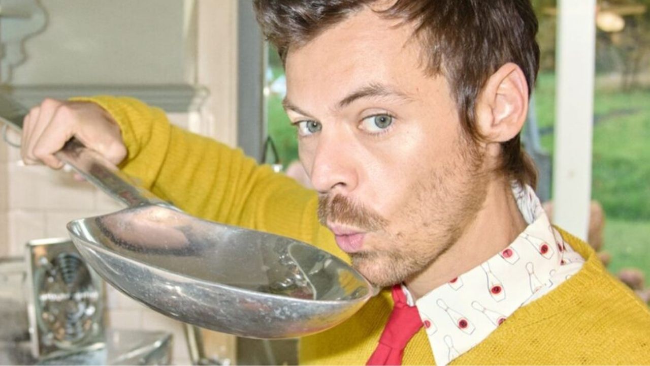 Harry Styles com suéter amarelo sopra uma colher de metal, em ambiente interno.