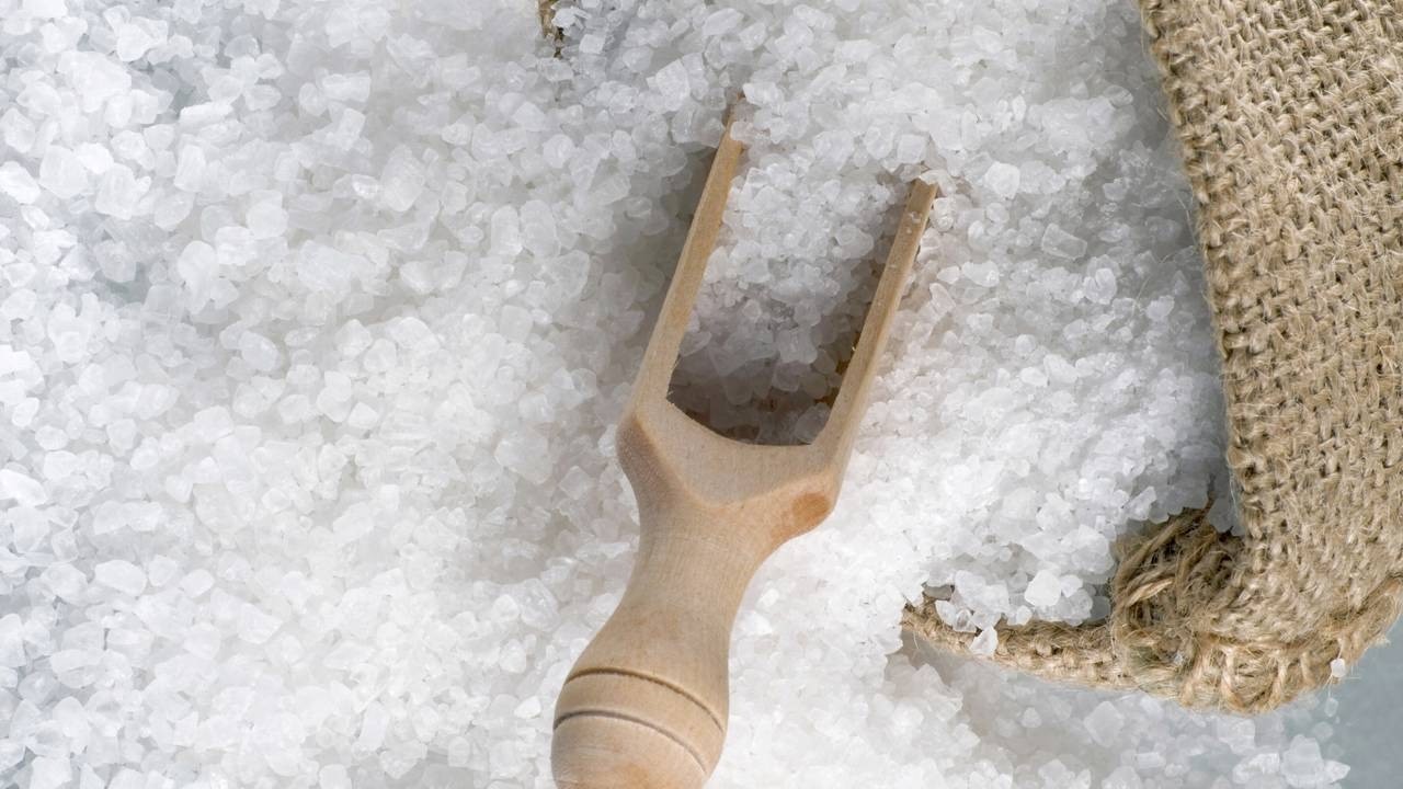 Close-up de cristais de sal branco granulado com uma pequena pá de madeira e um saco de estopa.