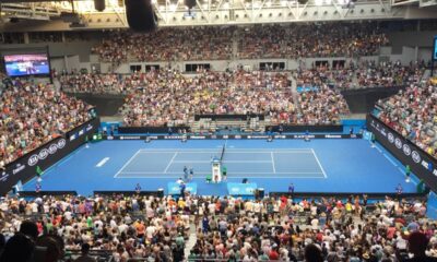 Quadra de tênis azul com jogadores, árbitro, telões e público lotado em estádio indoor. Logos: Kia, Mastercard, Hisense.