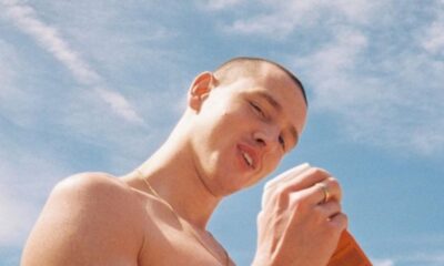 Harris Dickinson, homem sem camisa, cabelo raspado, sorrindo, segurando objeto contra céu azul.