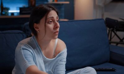 Mulher com expressão compenetrada assiste à TV em sofá azul, à noite, em sala escura.