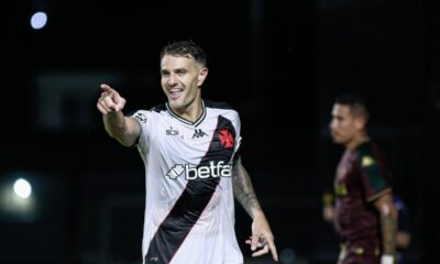 Jogador Vegetti do Vasco sorri e aponta em campo com camisa listrada e patrocinador.