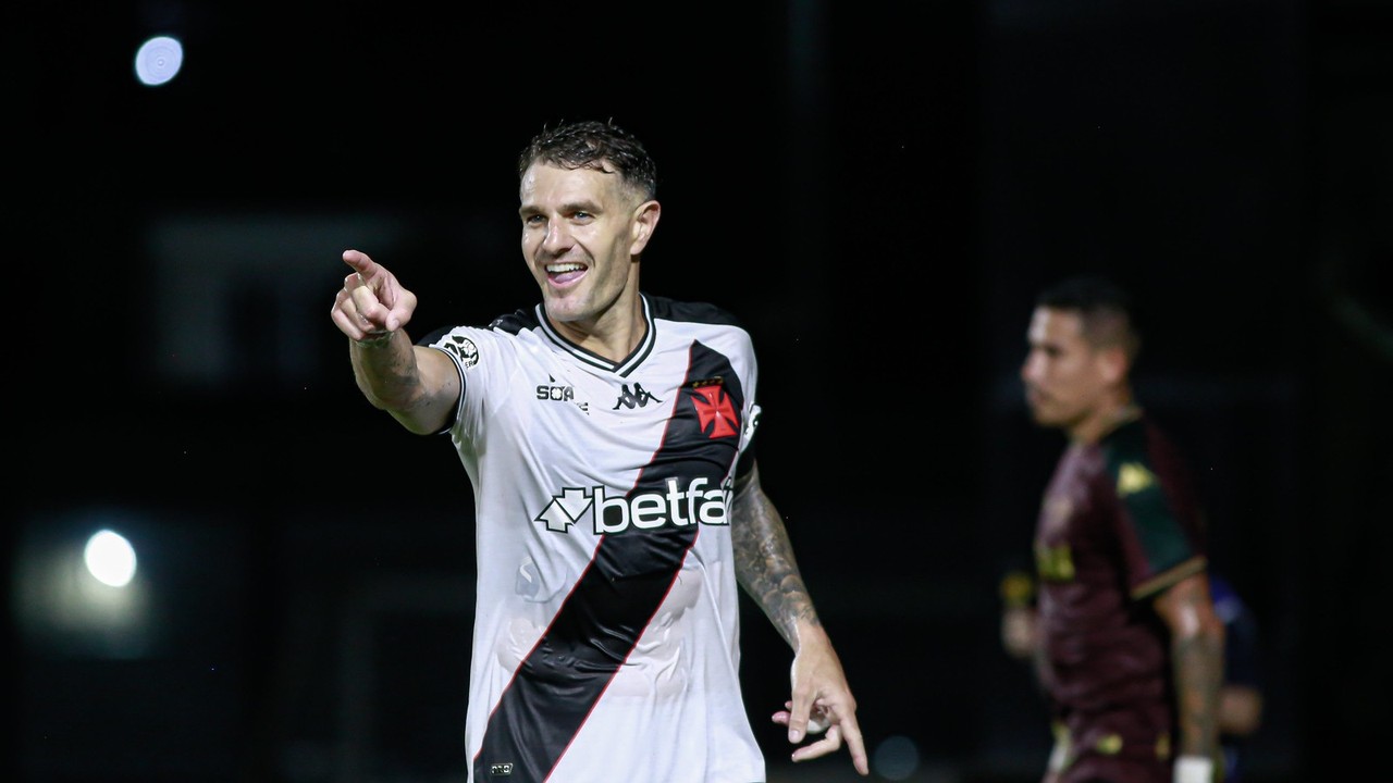 Jogador Vegetti do Vasco sorri e aponta em campo com camisa listrada e patrocinador.