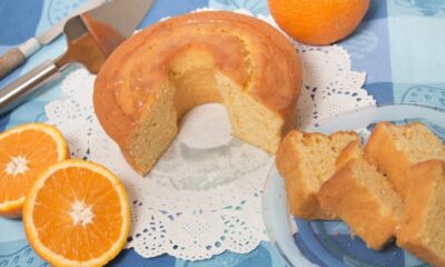 Bolo de laranja redondo com fatia cortada, laranjas frescas e fatias de bolo em prato azul.