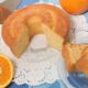 Bolo de laranja redondo com fatia cortada, laranjas frescas e fatias de bolo em prato azul.