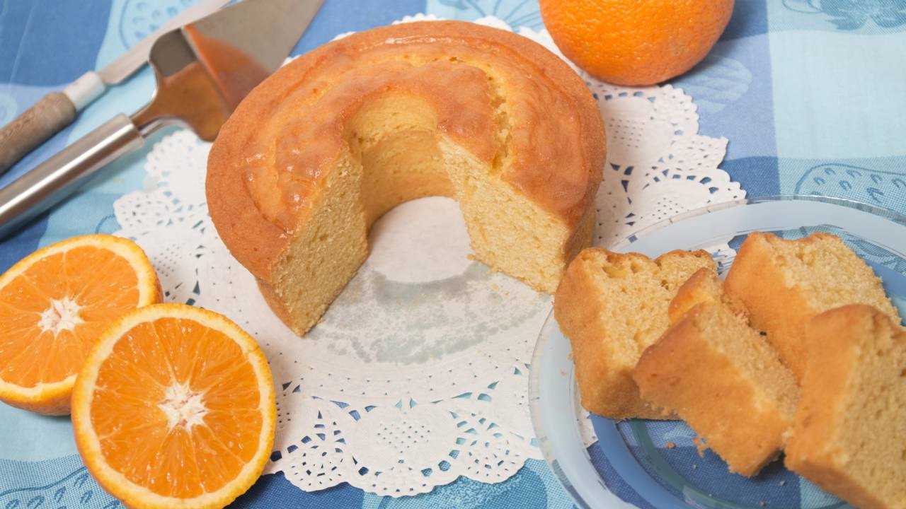 Bolo de laranja redondo com fatia cortada, laranjas frescas e fatias de bolo em prato azul.