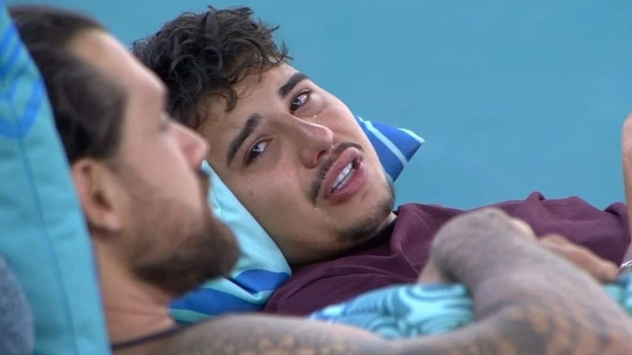 Homem jovem deitado em travesseiro azul, chorando visivelmente, com outro homem ao lado.