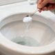 Mão despejando sal granulado branco de uma colher de metal em um vaso sanitário branco.
