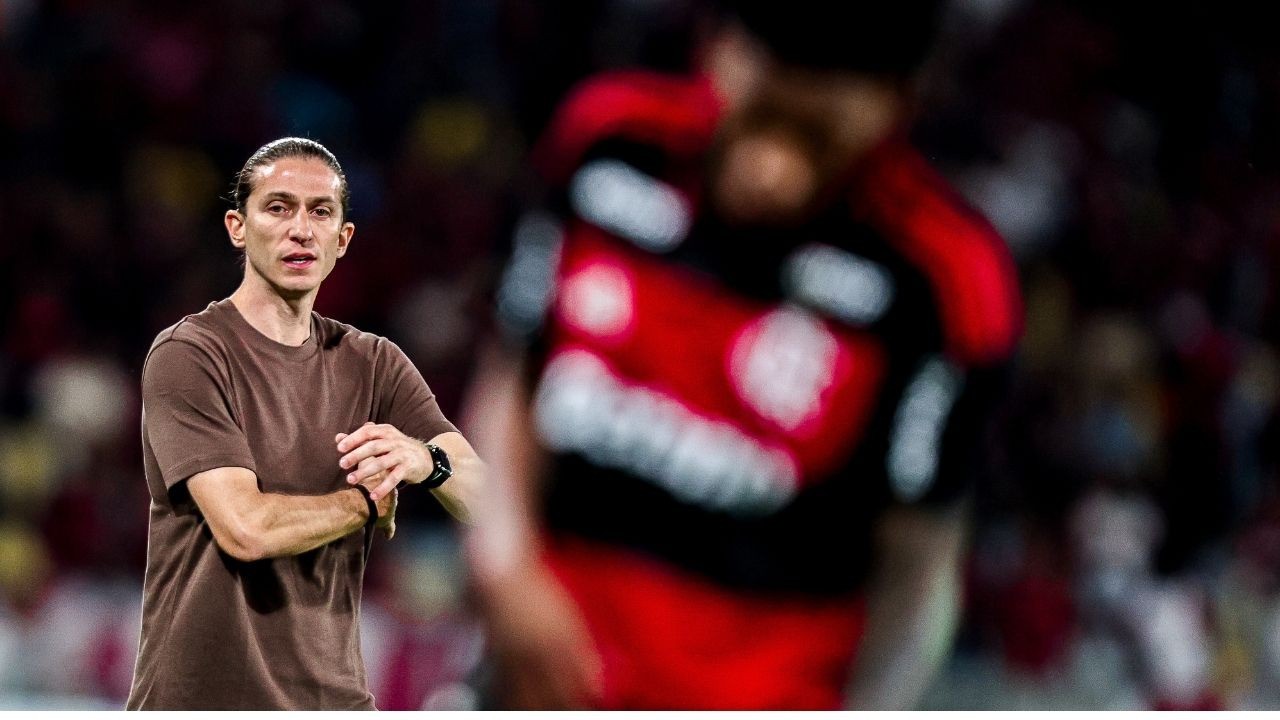 Homem de cabelo comprido, camiseta marrom, braços cruzados, em pé em estádio borrado.