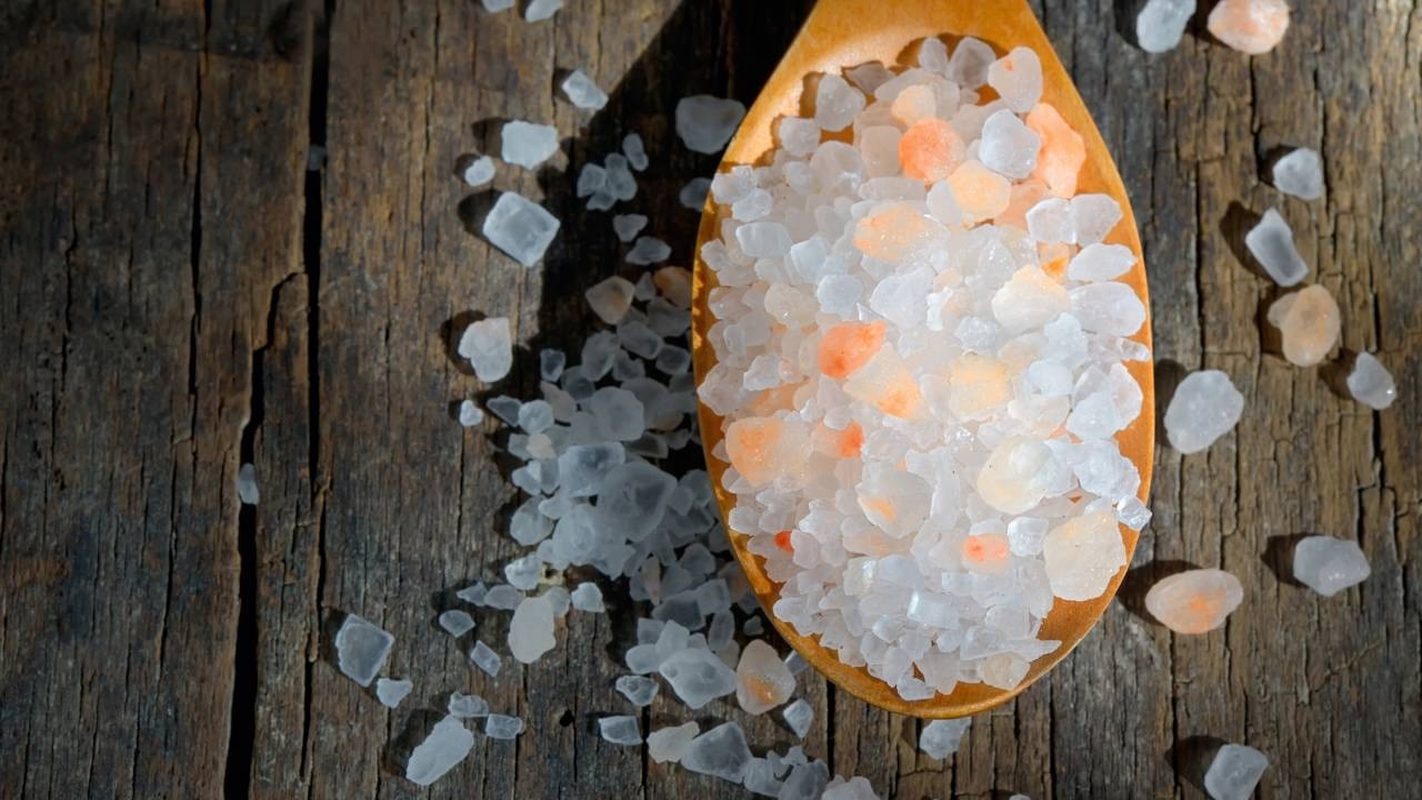 Colher de madeira com sal grosso branco e rosa sobre tábuas de madeira rústicas, sob luz.