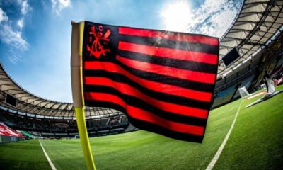 Bandeira preta e vermelha do Flamengo em campo de futebol, com estádio ao fundo e sol.
