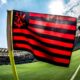 Bandeira preta e vermelha do Flamengo em campo de futebol, com estádio ao fundo e sol.