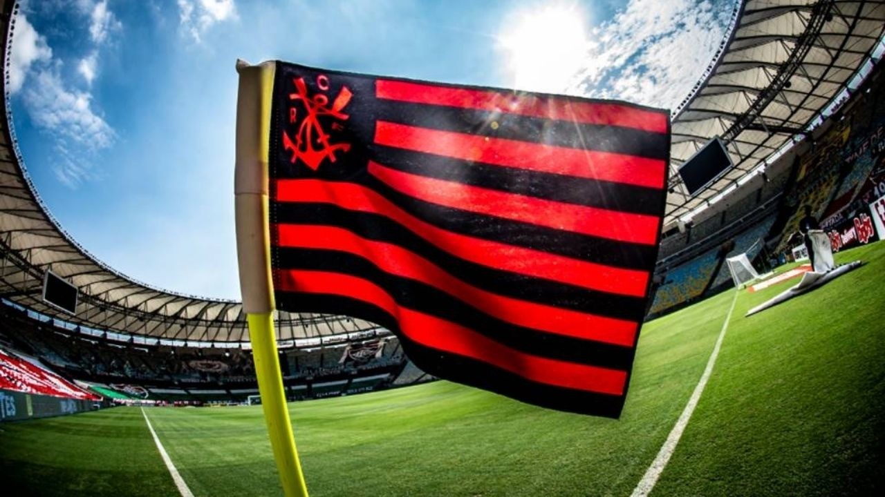 Bandeira preta e vermelha do Flamengo em campo de futebol, com estádio ao fundo e sol.