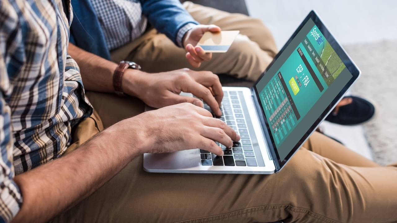 Mãos digitam em laptop exibindo site "SportsBet"; outra mão segura um cartão de crédito.