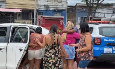 Pessoas abraçadas perto de um carro branco e uma viatura policial azul em uma rua.
