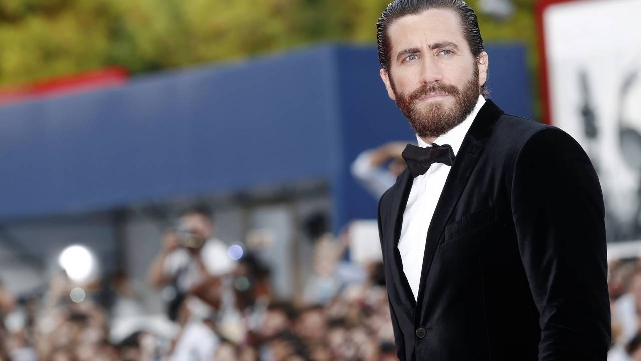 Jake Gyllenhaal, barba, cabelo penteado, veste terno de veludo preto e gravata borboleta em evento.