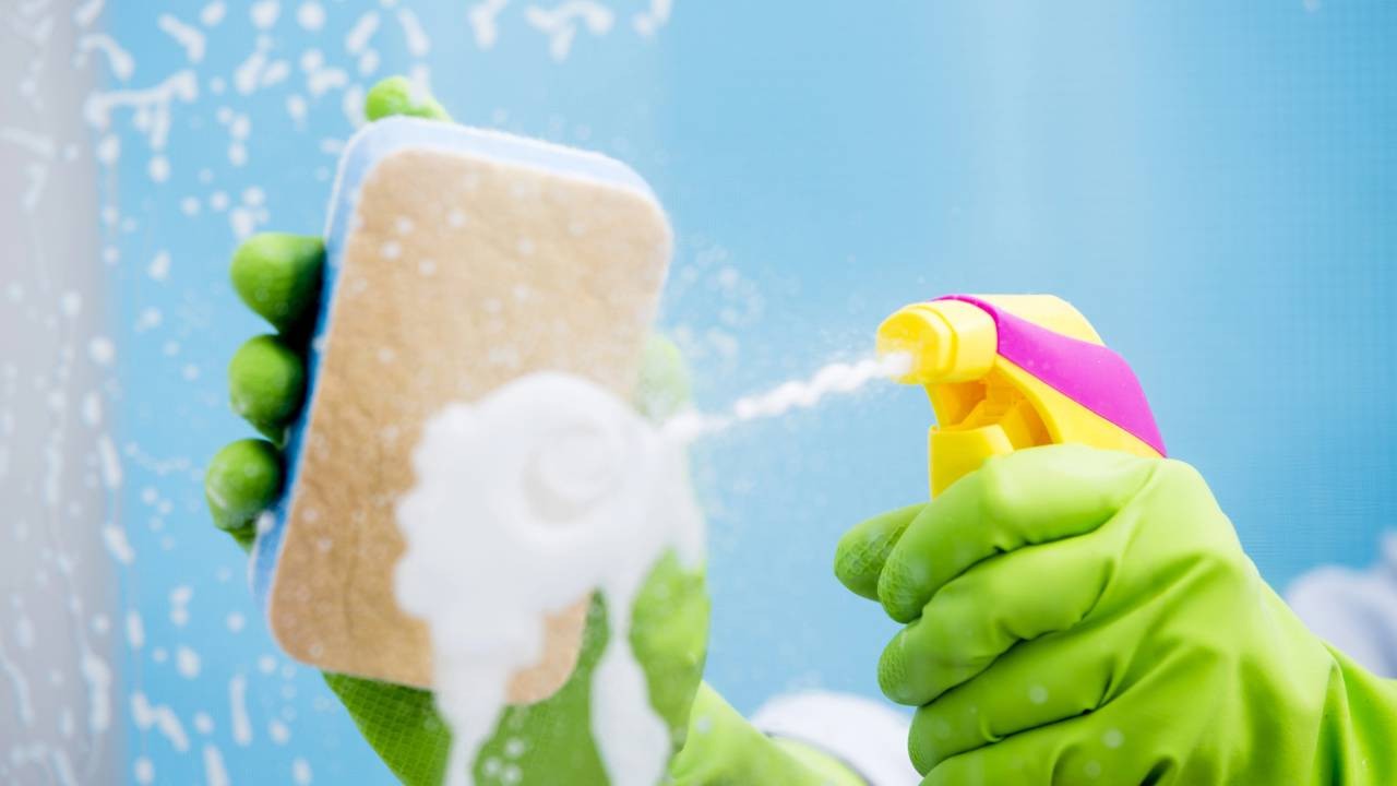 Mãos com luvas verdes seguram esponja e borrifador amarelo, limpando superfície azul com spray.
