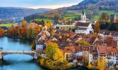 Vista aérea de cidade suíça com rio, ponte, casas coloridas, igreja e colinas arborizadas no outono.